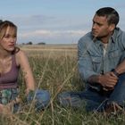 Zwei Menschen sitzen auf einer Wiese. Beide tragen lässige Kleidung. Der Himmel ist leicht bewölkt.