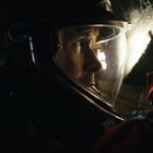 Ein Astronaut mit Helm sieht konzentriert nach rechts. Der Raum ist dunkel, mit Lichtreflexen auf dem Helmvisier.