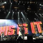 Text "THIS IS IT" in großen, beleuchteten Buchstaben. Menschen performen auf einer Bühne mit hellen Scheinwerfern.