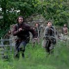 Zwei Menschen mit Bogen rennen durch eine grüne, bewaldete Landschaft. Im Hintergrund sind weitere Personen zu sehen.