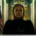 Eine Person mit ernster Miene steht vor zwei Flaggen und einem großen Siegel im Hintergrund.