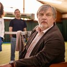 Eine Frau und ein Mann stehen hinter einem älteren Mann an einem Tenniscourt. Der ältere Mann trägt einen Anzug.