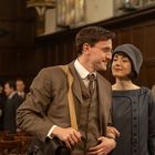 Ein Mann und eine Frau in eleganter Kleidung lächeln sich in einem Raum mit Holzwänden an, während andere Menschen im Hintergrund stehen.