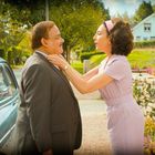 Ein Mann in Anzug und eine Frau in lila Kleid lächeln sich an. Sie richtet seine Krawatte. Im Hintergrund ein altes Auto.