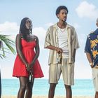Vier Personen stehen am Strand, entspannt gekleidet, mit Getränken in der Hand, vor blauem Meer und Himmel.