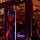 In einem Käfig liegt jemand in schwarzem Latexanzug und Gasmaske. Die Szene ist in Rotlicht getaucht.