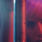 Das Bild zeigt eine Person mit lila Haaren, die nah an einer Stange steht. Beleuchtung in Blau und Rot.