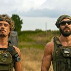 Zwei Männer in militärischer Kleidung im Freien, einer mit Zigarre, der andere mit Sonnenbrille.