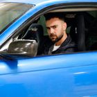 Ein Mann sitzt in einem blauen Auto. Auf dem Sitz steht "bmwrec". Er hat kurzes Haar und einen Bart, schaut ernst.