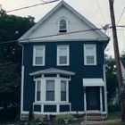 Ein zweistöckiges, blaues Haus mit weißen Akzenten. Davor sind Bäume, und ein Auto steht rechts neben einem kleineren Haus.