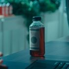 Auf einem Tisch steht eine Flasche mit dem Etikett "Mother's Secret Kombucha". Im Hintergrund sieht man unscharfe Pflanzen.