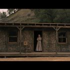 Eine Frau steht auf der Veranda einer rustikalen Holzhütte, umgeben von Natur und Bäumen.