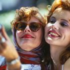 Zwei Frauen mit roten Lippen, lächelnd, machen ein Selfie. Eine trägt eine Sonnenbrille und ein buntes Kopftuch.