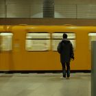Ein U-Bahn-Zug in Gelb rauscht vorbei, darauf der Text "Bundesta". Eine Person steht auf dem Bahnsteig. Uhr und Anzeige darüber.