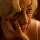 Eine ältere Frau mit roten Nägeln stützt nachdenklich ihr Gesicht mit der Hand. Sie hat graue Haare und trägt ein weißes Kleid.