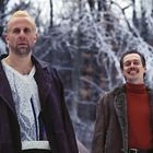 Zwei Männer stehen im Schnee. Links ein Mann in Lederjacke, rechts ein Mann mit Fellmantel. Hintergrund: vereiste Bäume.