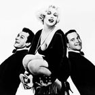 Ein Mann und eine Frau in schwarzer Kleidung, beide mit freudigem Ausdruck. Die Frau hält eine Ukulele, zwischen zwei Männern.