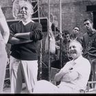 Keine sichtbaren Texte. Eine Gruppe von Männern in entspannter Atmosphäre; einer sitzt und lacht, andere stehen.