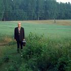 Ein Mann in einem dunklen Anzug steht auf einer grünen Wiese mit Wald im Hintergrund, er hält einen Koffer.