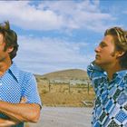 Kein Text vorhanden. Zwei Männer in gemusterten Hemden stehen draußen, entspannte Haltung, blauer Himmel, Hügel im Hintergrund.