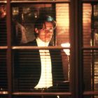 Eine Person in einem hell beleuchteten Raum schaut durch ein Fenster mit Jalousien. Schatten und Lichter erzeugen Kontraste.