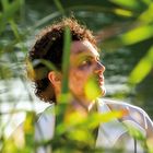 Ein Mann mit lockigem Haar blickt nach oben, umgeben von grünen Pflanzen am Wasser.