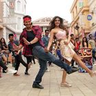 Menschen tanzen in einer lebhaften Straßenkulisse mit bunten Graffiti-Tonnen. Zwei Personen stehen im Vordergrund.