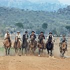 Sieben Reiter in Cowboy-Outfits galoppieren durch eine trockene, staubige Landschaft mit Hügeln im Hintergrund.