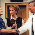 Ein Mann in einem weißen Kittel schüttelt die Hand einer lächelnden Frau. Eine ernst blickende Frau schaut zu.