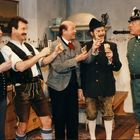 Fünf Männer in traditioneller bayerischer Kleidung, jeder hält Spielkarten, sprechen mit einem Polizisten in Uniform.