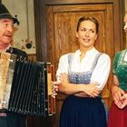Ein Mann spielt Akkordeon, zwei Frauen in Dirndl lächeln. Traditionelle bayerische Kleidung, Holzwand im Hintergrund.