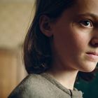 Es gibt keinen sichtbaren Text. Ein junges Mädchen blickt ernst zur Seite, im Hintergrund ein verschwommener Raum.