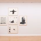 Eine Person betrachtet sechs gerahmte schwarz-weiße Kunstwerke an einer weißen Galeriewand. Ein Notausgangsschild ist rechts.