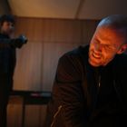 Ein Mann kniet mit schmerzhafter Miene im Vordergrund. Eine Person im Hintergrund zielt mit einer Pistole auf ihn.