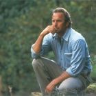Ein nachdenklicher Mann in einem blauen Hemd hockt im Grünen. Er legt eine Hand ans Kinn.