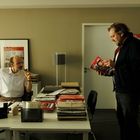 Eine Person sitzt in einem Büro, während eine andere ein Magazin namens "fakt." hält. Der Raum ist mit Akten gefüllt.