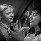 Zwei Personen in einem Gespräch. Der Mann lächelt, während die Frau nachdenklich wirkt und etwas hält. Schwarz-weiß.