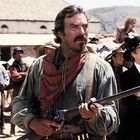 Ich kann keine Texte erkennen. Ein Mann im Western-Outfit hält ein Gewehr. Im Hintergrund eine Gruppe von Menschen.