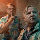 Ein Mann in bunter Hawaii-Hemd macht «Pssst»-Geste, ein anderer blickt nachdenklich weg. Orange Licht im Hintergrund.