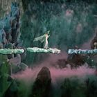 Eine Figur schreitet über schwebende Stege in einer mystischen Szenerie mit bläulich-grünen Felsen und Nebel.