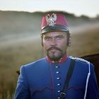 Ein Mann in einer Uniform mit roter Kopfbedeckung und einem silbernen Adleremblem steht auf einem Feld. Im Hintergrund ist Grasland.