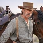 Ein Mann mit Hut und Bart steht mit entschlossenem Blick zwischen zwei Pferden, mit Satteln im Hintergrund.