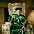 Ein Mann in einem grünen Gewand sitzt auf einem steinernen Thron mit asiatischen Mustern, trägt einen runden Hut.