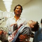 Text "EMERGENCY" an der Wand eines Krankenhausflurs. Eine Person trägt ein blutiges Kind, von einem Arzt begleitet.