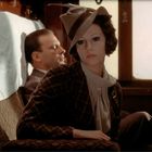 Eine Frau in eleganter Kleidung, die in einem Zug sitzt, schaut nachdenklich. Eine Blumenvase ist im Hintergrund.