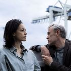 Eine Frau und ein Mann in einem Gespräch auf einem Boot, im Hintergrund unscharfe Hafenkräne unter bewölktem Himmel.