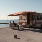 Ein Wohnmobil mit markanter Markise steht am Meer. Vor dem Fahrzeug sitzen Menschen auf Klappstühlen und entspannen.
