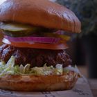 Ein saftiger Hamburger mit Salat, Tomate, Zwiebel, Gewürzgurke und Sauce auf einem Tablett in einem gemütlichen Restaurant.