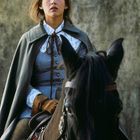 Keine Texte erkennbar. Eine Person in historischer Kleidung reitet auf einem schwarzen Pferd vor einem strukturierten Hintergrund.