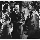 Drei Menschen stehen in einem eleganten Raum, zwei Männer im Anzug, eine Frau in Kleidung aus früheren Zeiten.